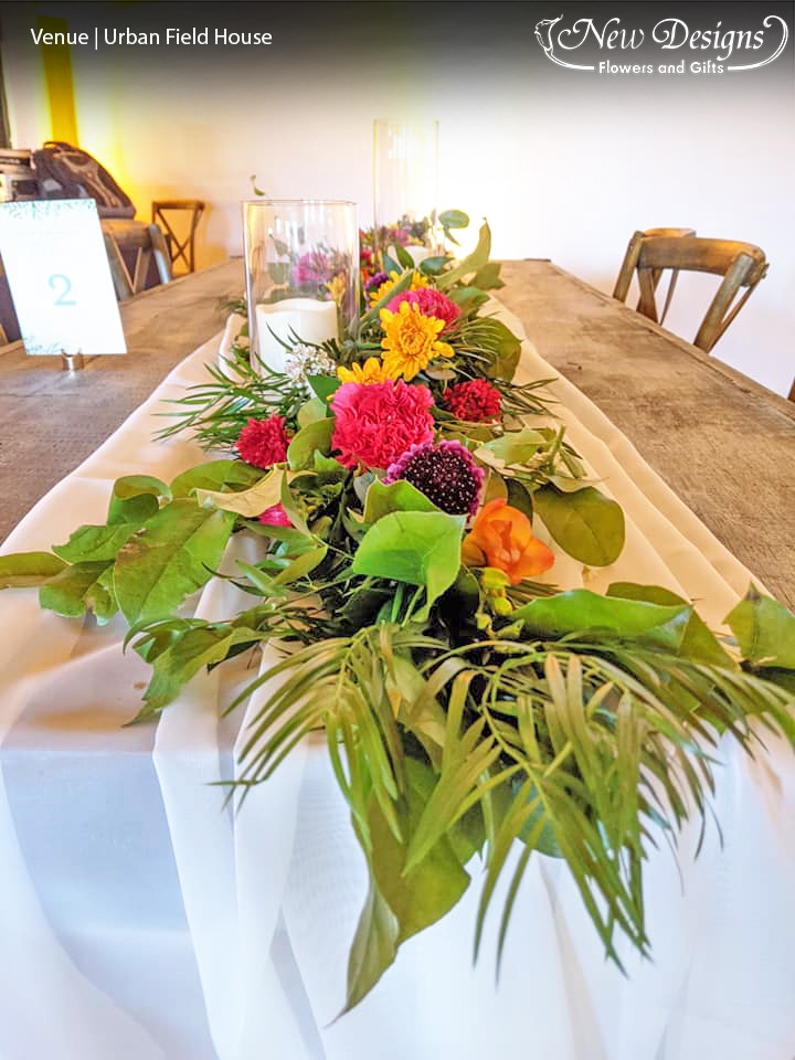 a bright garland of deep purples and orange exotic and wild flowers run atop spiky greens, along a white chiffon runner on top of a long wooden table